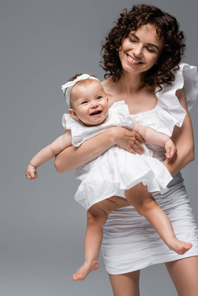 Woman holding baby girl in headband and white dress isolated on grey  - Фото, изображение