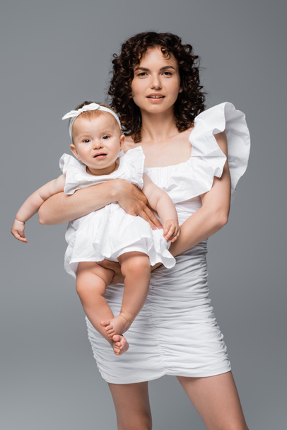 Trendy mother in dress looking at camera while holding baby girl isolated on grey  - Foto, imagen
