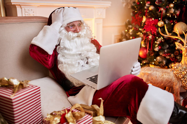 Mikulás ül az otthonában, és e-mailt olvas a laptopon a karácsonyi kérő vagy kívánságlista közelében a kandalló és a fa ajándékokkal. Szilveszter és Boldog Karácsonyt, Boldog Ünnepeket koncepció - Fotó, kép