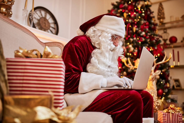 Der Weihnachtsmann sitzt zu Hause und liest E-Mails auf dem Laptop mit Weihnachtsanfragen oder Wunschzetteln neben dem Kamin und dem Baum mit Geschenken. Neues Jahr und frohe Weihnachten, frohe Feiertage - Foto, Bild