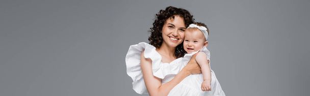 Happy mother in white dress hugging baby daughter isolated on grey, banner  - Fotoğraf, Görsel