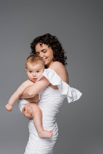 Happy woman in stylish white dress hugging infant child isolated on grey  - Фото, зображення