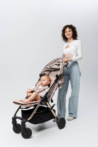 Full length of smiling mother looking at camera near baby in stroller on grey background  - Foto, Bild