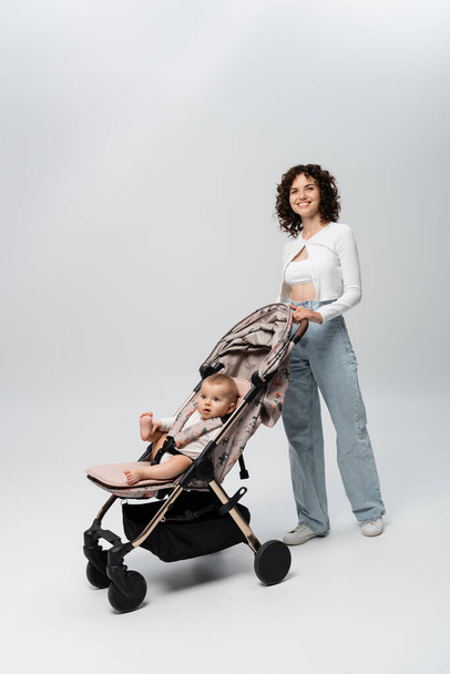 Curly parent standing near infant daughter in stroller on grey background  - Foto, afbeelding