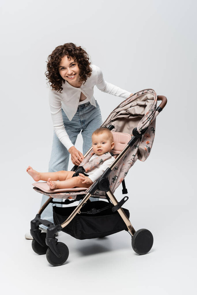 Happy mother looking at camera near baby girl in stroller on grey background  - Foto, imagen