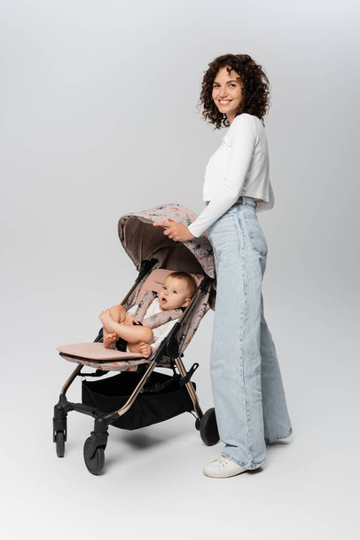 Кудрявая мать в джинсах стоит рядом с маленькой девочкой в коляске на сером фоне  - Фото, изображение