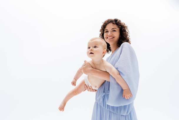 Cheerful woman in blue dress looking at camera while holding infant girl isolated on white  - Foto, Imagen