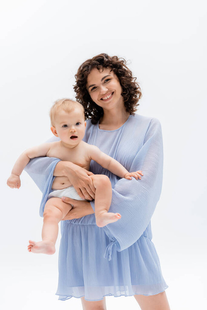 Mujer rizada positiva en vestido sosteniendo al bebé en bragas aisladas en blanco  - Foto, Imagen
