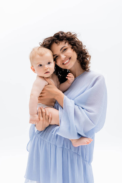 gelukkig ouder in blauw jurk kijken naar camera en aanraken baby dochter geïsoleerd op wit  - Foto, afbeelding