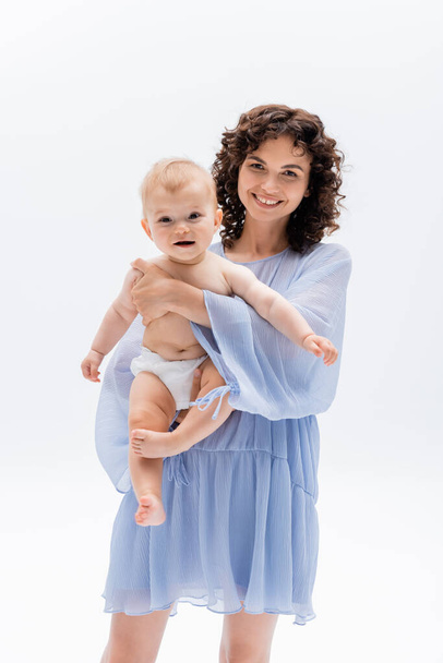 Woman in blue dress hugging baby in panties and looking at camera isolated on white  - Foto, afbeelding