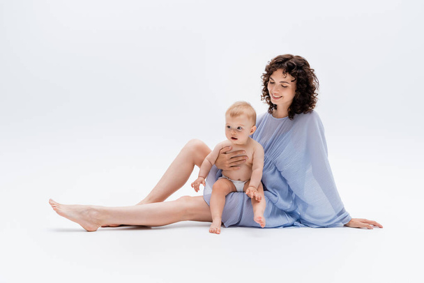 Curly woman in dress looking at infant daughter while sitting on white background  - Foto, Imagem