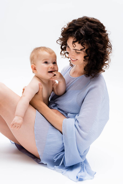 Mosolygó anya ruhában nézi elégedett baba lánya, miközben ül a fehér háttér  - Fotó, kép