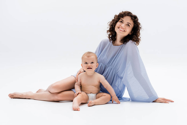 Smiling parent in dress touching baby daughter while sitting on white background  - Foto, Imagem