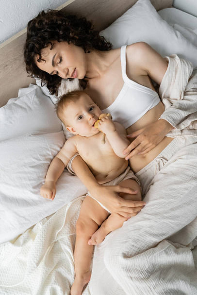 Bovenaanzicht van moeder kijken naar baby meisje met houten speelgoed terwijl liggend op bed  - Foto, afbeelding