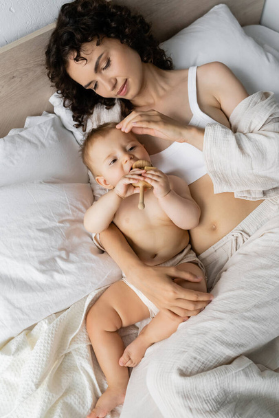 Vista superior de la mamá morena en pijama tocando a la hija con juguete en la cama  - Foto, imagen