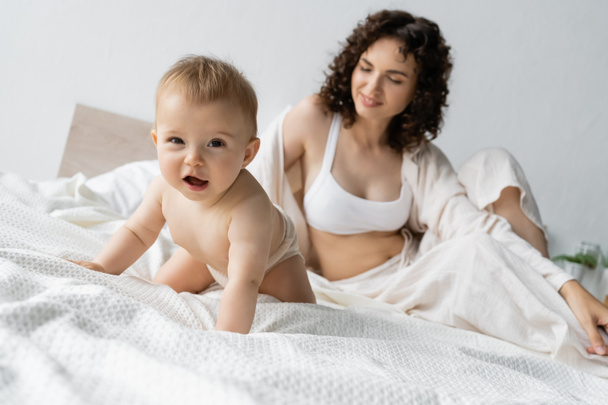 Fröhliches Babymädchen blickt im Schlafanzug auf dem Bett in die Kamera  - Foto, Bild