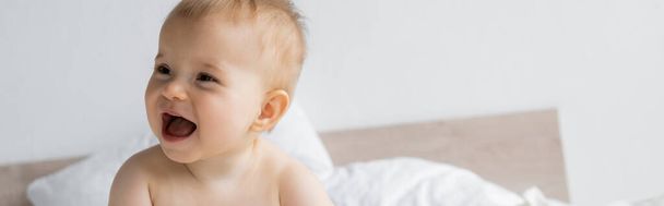 Cheerful baby girl looking away on blurred bed at home, banner  - Фото, изображение
