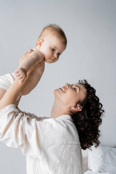 Curly woman smiling while lifting baby girl in bedroom in morning  - Photo, Image