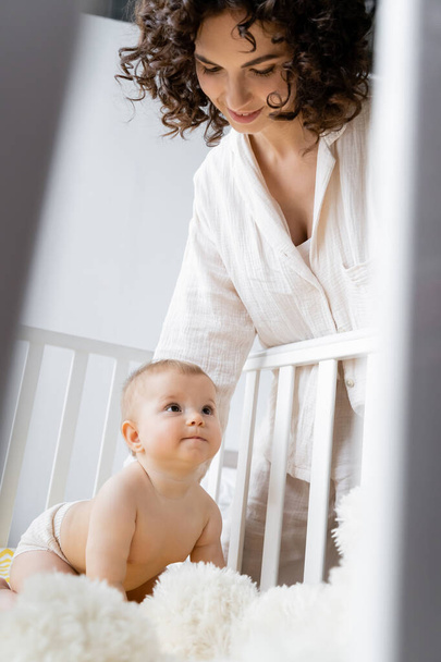 Hymyilevä nainen katselee vauva tyttö lähellä hämärtynyt pehmeä lelu pinnasänky  - Valokuva, kuva