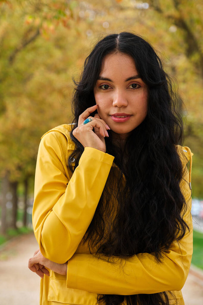 若いですラテン女の子身に着けていますで黄色のレインコート姿で見ますカメラでストリートで秋. - 写真・画像