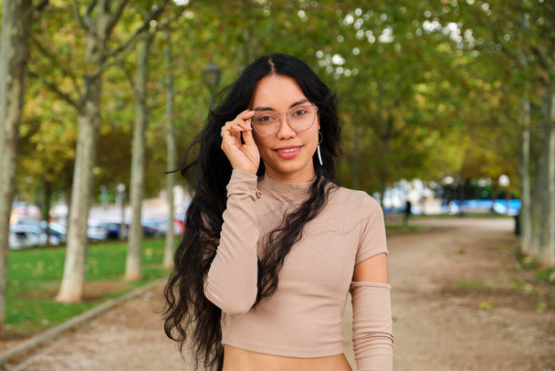 Fiatal latin lány fogja a szemüvegét és mosolyogva néz ar kamera az utcán ősszel. - Fotó, kép