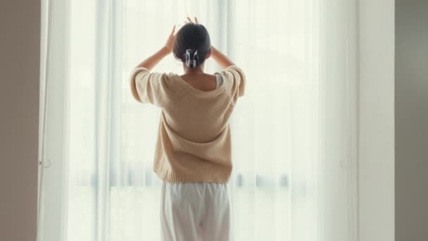 Glückliche asiatische Mädchen mit weißen cremefarbenen Pyjamas offenen weißen Vorhang am Fenster nach draußen fühlen sich frisch mit friedlichem Morgen und frischer Luft am Wochenende im Schlafzimmer zu Hause. Weibliches Lebensstil-Konzept. - Filmmaterial, Video