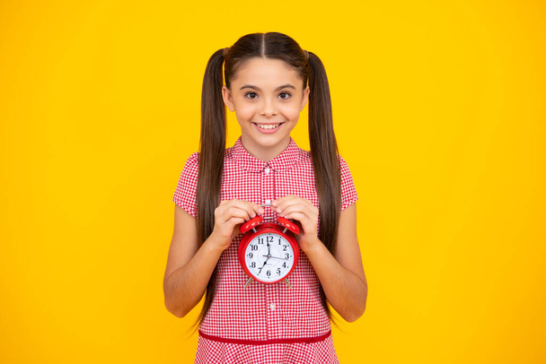 十代の子供は黄色のスタジオの背景に隔離された時計を保持する。目覚まし時計を持つ10代の子供は時間を示す。幸せなティーン,正と笑顔感情のティーン女の子 - 写真・画像