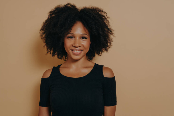 Портрет веселої усміхненої красивої темношкірої дівчини в чорній сорочці, що стоїть ізольовано на темно-бежевому фоні стіни з копіювальним простором для реклами. Концепція молодості та щастя
 - Фото, зображення
