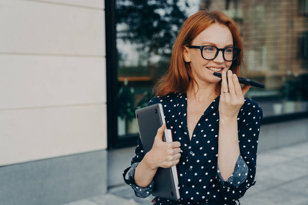 Piękna rudowłosa kobieta biznesowa stoi na zewnątrz z telefonem komórkowym i nagrywa wiadomość audio lub smartfon, rozmawia z wirtualnym cyfrowym asystentem głosowym w drodze do biura, kobieta korzystająca z głośnomówiącego - Zdjęcie, obraz