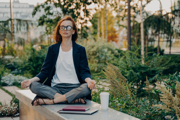 Full length shot of relaxed ginger woman meditates sits in lotus pose wears formal clothes keeps legs crossed breathes deeply spends free time in park surrounded by takeaway coffee tablet and notepad - Photo, Image