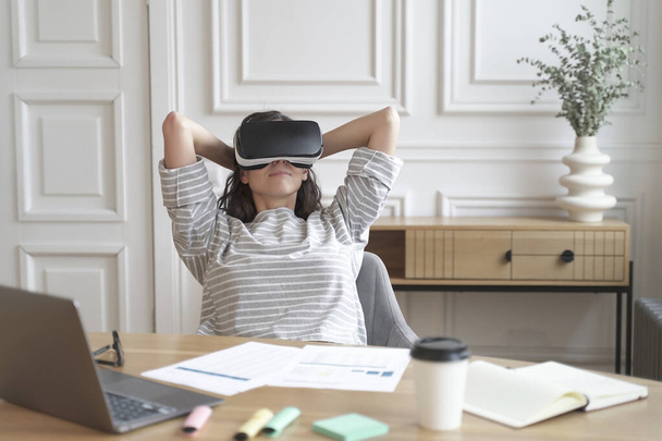 Mladá ráda uvolněná žena kancelářská pracovnice ve virtuální realitě brýle na pracovišti s notebookem, usmívající se žena zaměstnanec relaxace ve VR helmě během přestávky v práci - Fotografie, Obrázek