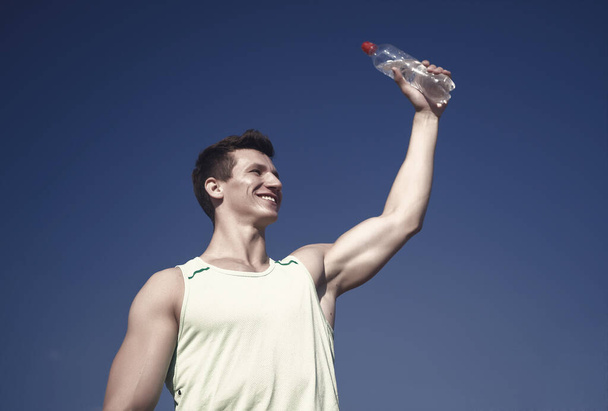 Athlete smiling in green tshirt on blue sky. Man with water bottle in muscular hands. Healthy lifestyle and health. Sport and fitness concept. Bodybuilder with muscles biceps, triceps on sunny day. - Fotografie, Obrázek