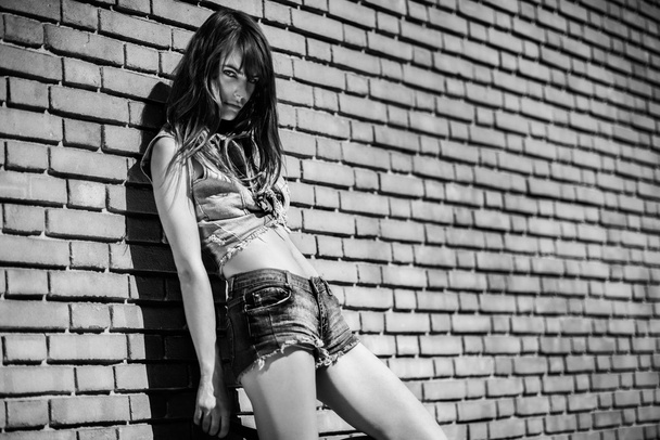 Beautiful young brunette in jeans clothes posing outdoor near red brick wall - Fotó, kép
