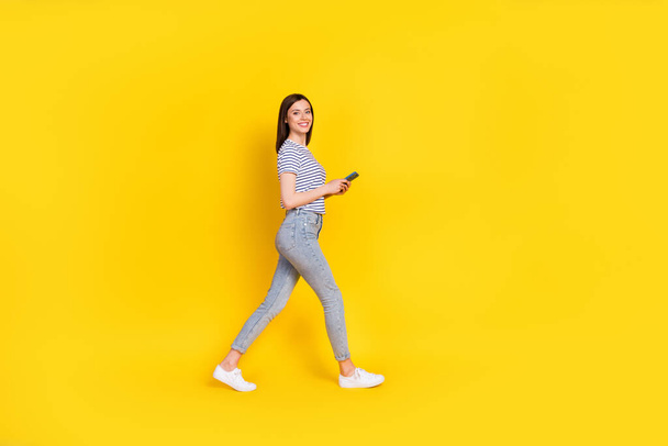 Full size profile photo of satisfied lady walking empty space arm hold use modern device gadget isolated on yellow color background. - Fotó, kép