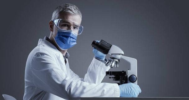 Medical scientist wearing a face mask and working in the laboratory with a microscope, he is looking at camera - Foto, Bild