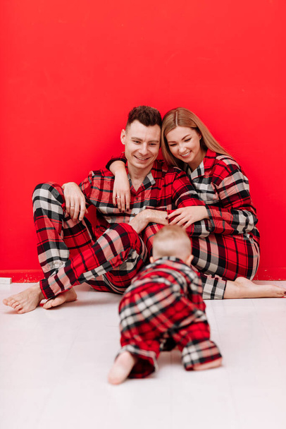 Happy family is having fun together. Smiling young mom, dad and baby son in red checked pyjamas sitting on red background. Mothers day. fathers day, baby day. Christmas. Valentines day. - 写真・画像
