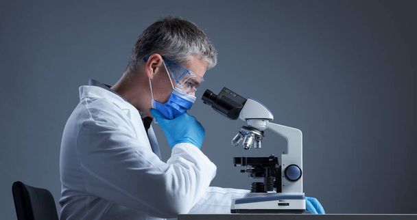 Scientist with face mask examining a sample under the microscope and thinking, scientific research concept, blank copy space - Foto, immagini