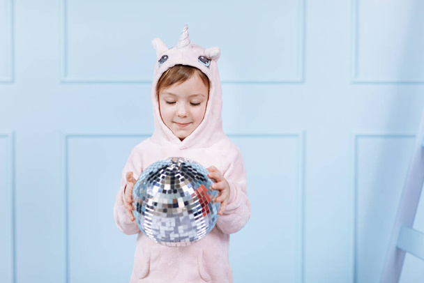 Sonriendo hermosa niña en traje rosa unicornio es sonreír y divertirse con la bola disco en un fondo de pared azul. feliz infancia - Foto, Imagen