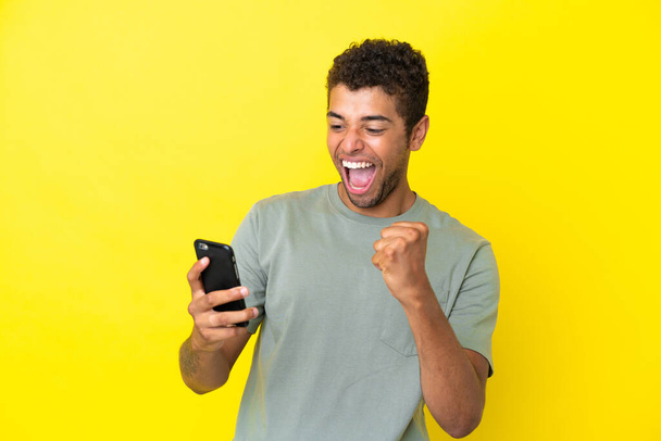 Joven brasileño guapo aislado sobre fondo amarillo con teléfono en posición de victoria - Foto, Imagen