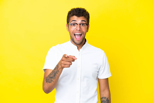 Young handsome Brazilian man isolated on yellow background surprised and pointing front - Φωτογραφία, εικόνα