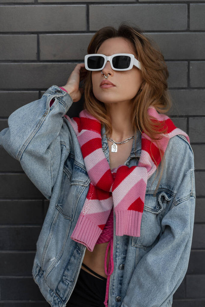 Modieuze trendy jonge mooie roodharige vrouwelijke model hipster met witte modezonnebril in blauwe jeans en roze sweatshirt staat en poseert in de buurt van zwarte bakstenen muur in de stad. Urban vrouwelijke stijl look - Foto, afbeelding