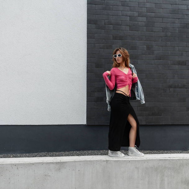 Μοντέρνο hipster όμορφη γυναίκα σε casual denim street outfit με τζιν, φούστα και παπούτσια περπατά κοντά σε ένα σύγχρονο τούβλο μαύρο άσπρο κτίριο - Φωτογραφία, εικόνα