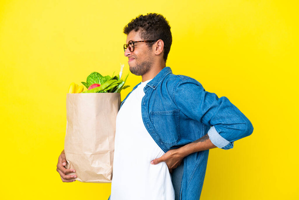 黄色の背景に孤立した食料品の買い物袋を持っている若いブラジル人男性は、努力をしたために腰痛に苦しんでいます - 写真・画像