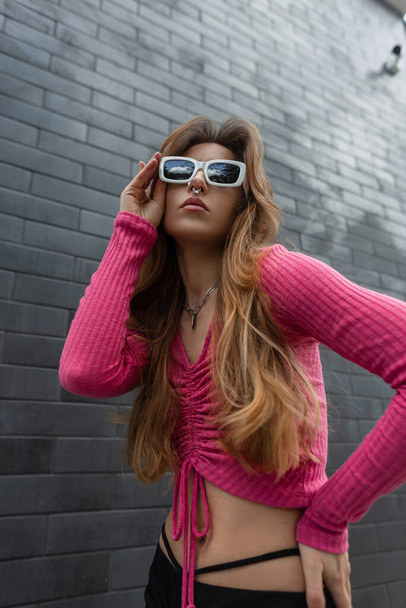 Cool jonge stijlvolle roodharige hippe vrouw model met mode witte zonnebril in een trendy roze gewas top en rok poseert in de buurt van een zwarte baksteen muur op de straat - Foto, afbeelding