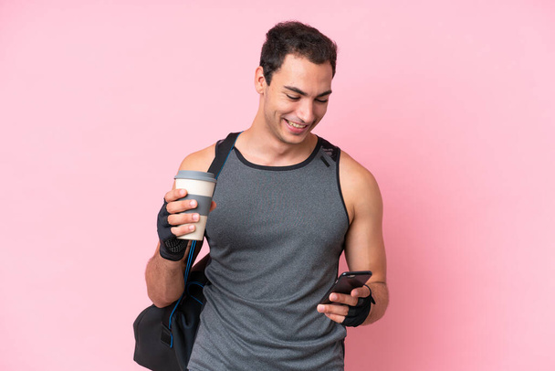 Νεαρός αθλητής καυκάσιος άνδρας με αθλητική τσάντα απομονωμένη σε ροζ φόντο κρατώντας καφέ για να πάρει μακριά και ένα κινητό - Φωτογραφία, εικόνα