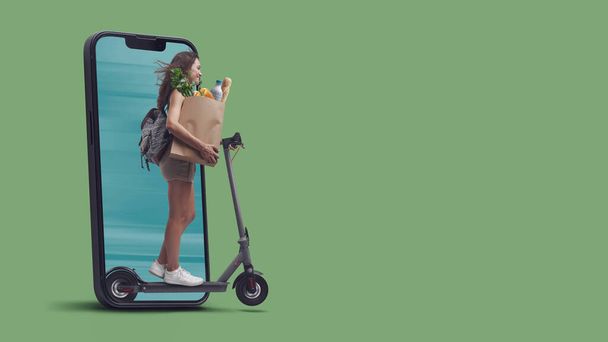 Donna in possesso di un sacchetto di carta con generi alimentari e in sella a uno scooter elettrico, sta uscendo da uno schermo smartphone, concetto di spesa online - Foto, immagini