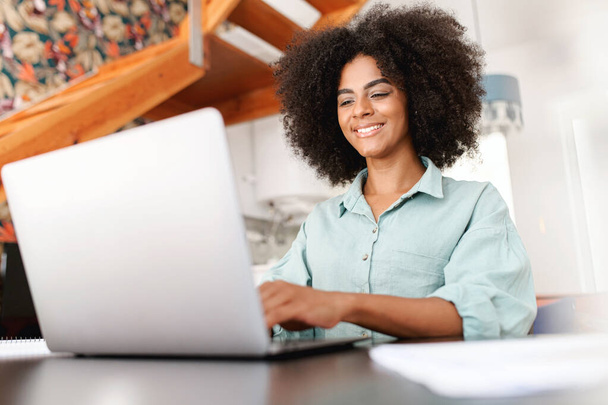Portrait of young african-american woman sitting at the desk with a laptop, typing an email, messaging friend, chatting with a pleasant smile. Female freelancer working remotely. Technologies concept - Fotografie, Obrázek