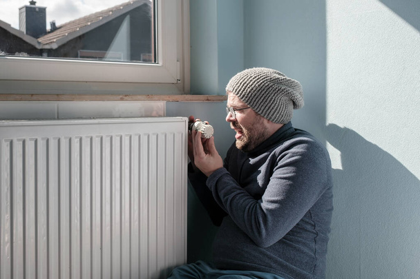 Annoyed man in a hat and a warm sweater, sits near the radiator and turns the thermostat to the minimum. Concept of crisis and increase in fuel costs. Preparing for a cold winter. - Photo, image
