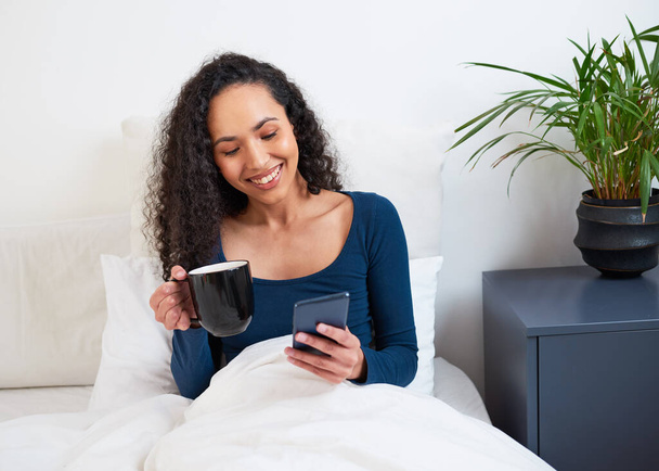 Mladá multietnická žena pije kávu v posteli, když se probouzí po telefonu. Kvalitní fotografie - Fotografie, Obrázek