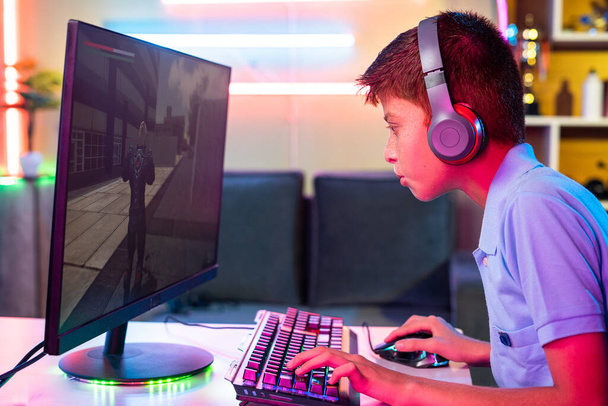 боковой вид ребенка-подростка с наушниками, играющего дома в онлайн видеоигру на компьютере - концепция соревновательной, развлекательной деятельности и игровой зависимости - Фото, изображение